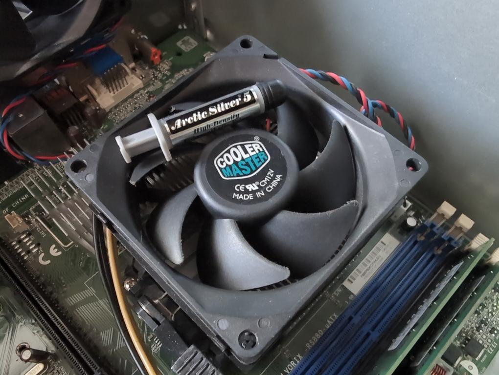 Photograph of a computer motherboard, focused on the CPU cooling fan. It has a small bottle of thermal paste on top of it.
